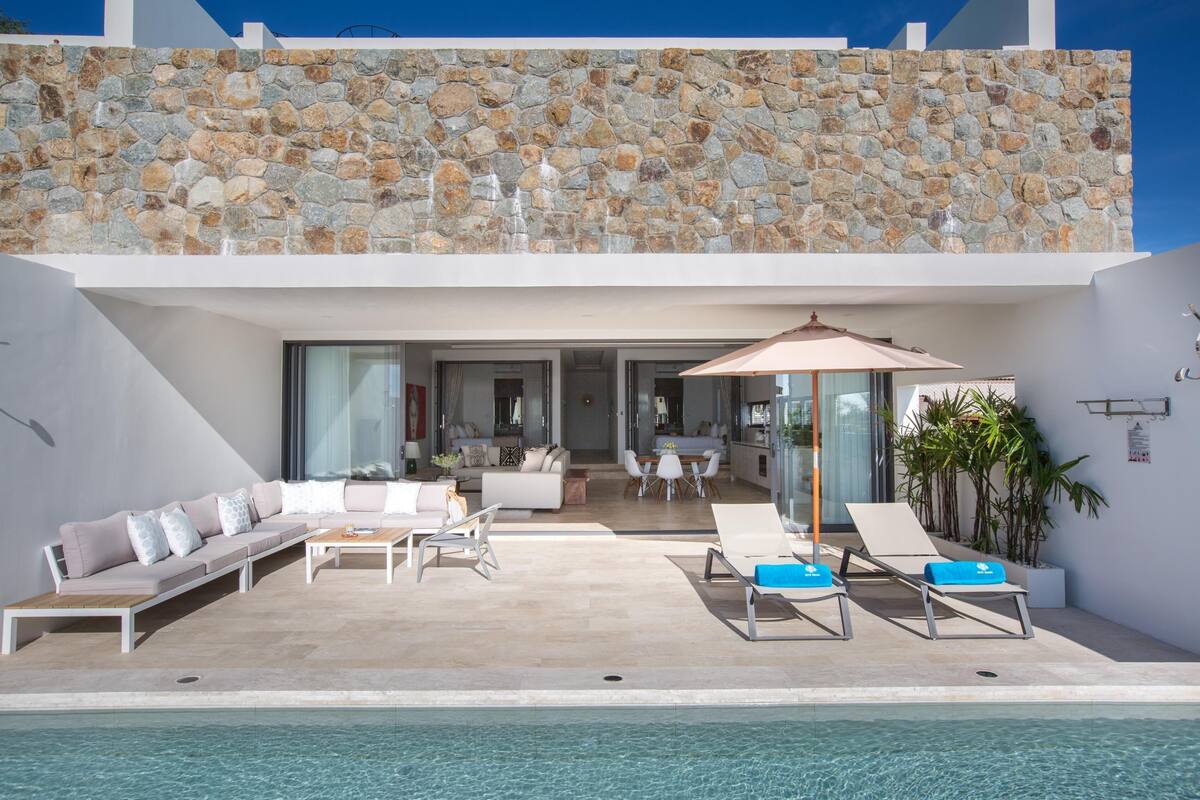 A modern outdoor pool area with a stone wall, a seating area with a sofa and chairs, and a dining area with a table and chairs.