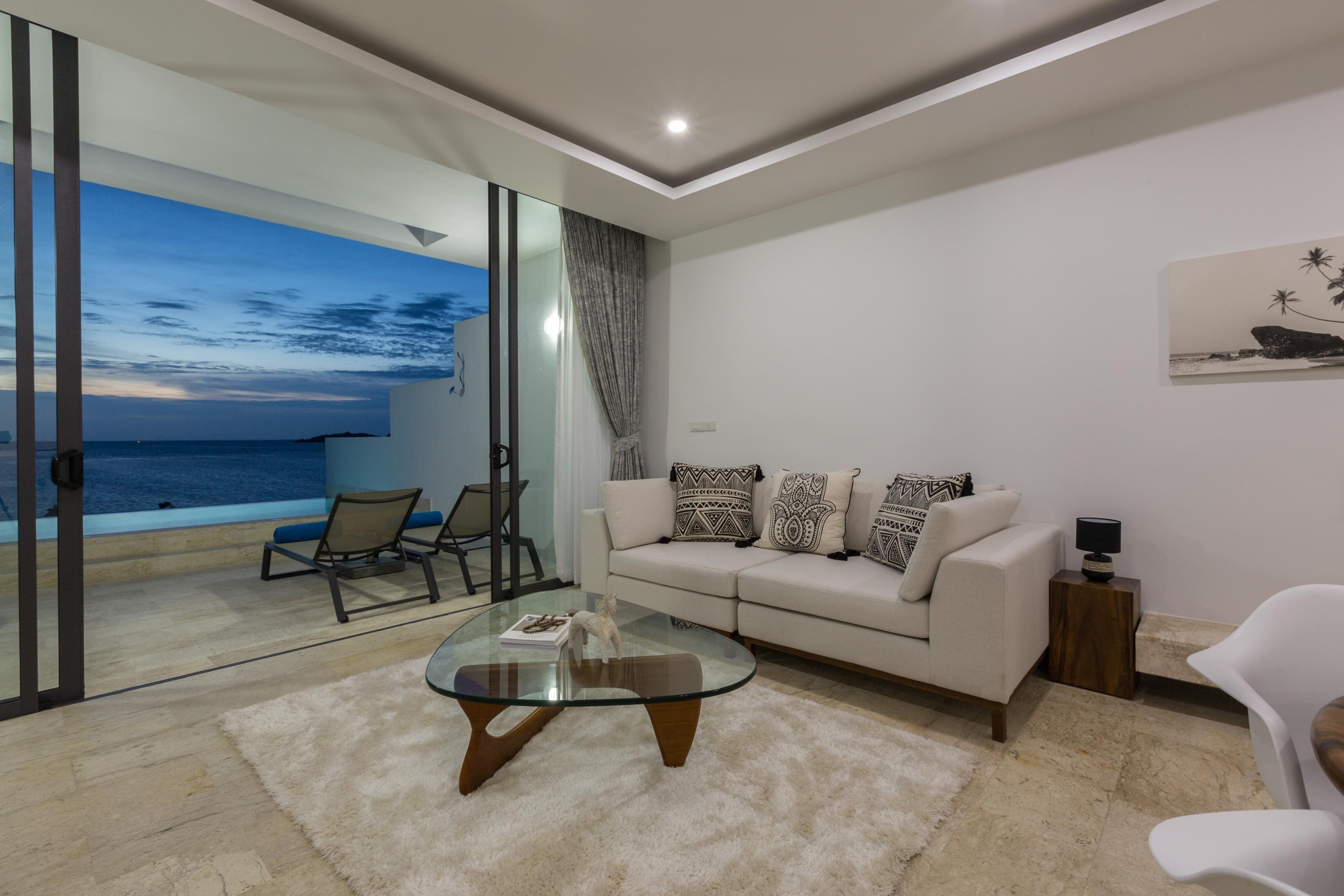 A modern living room with a glass coffee table, a sofa with patterned cushions, a white armchair, and a view of the ocean through large glass doors.