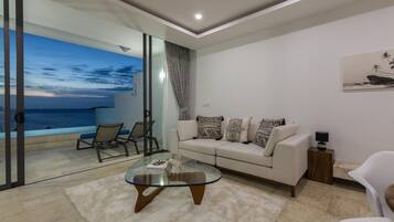 A modern living room with a glass coffee table, a sofa with patterned cushions, a white armchair, and a view of the ocean through large glass doors.