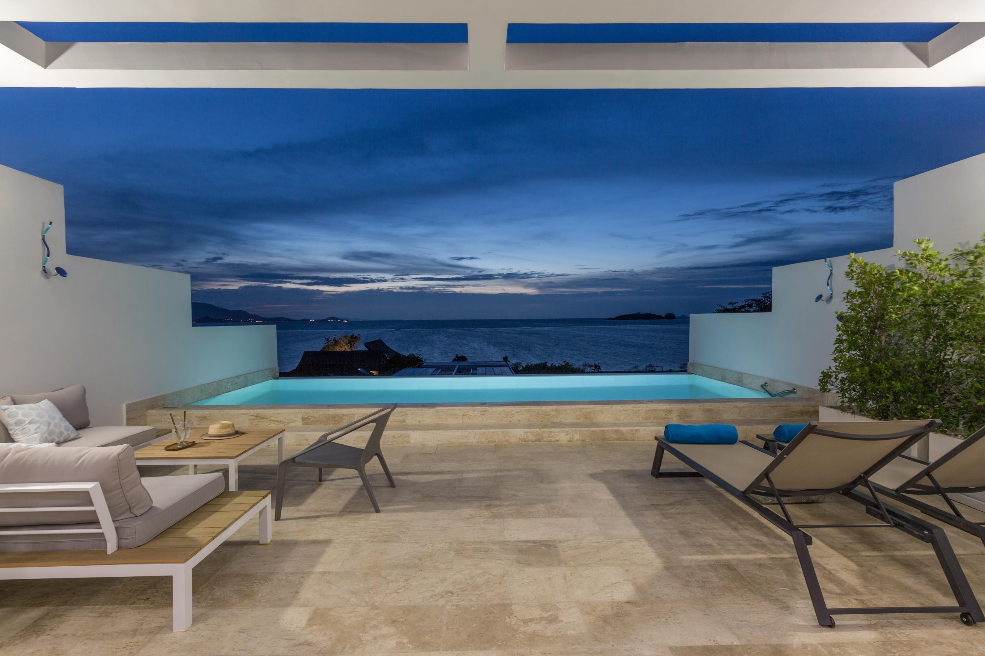 Un área de piscina moderna al aire libre con vista al mar, amueblada con sillones y un sofá.