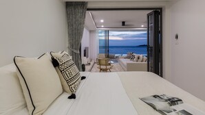 Un dormitorio moderno con una cama grande, vista al mar a través de una puerta abierta, un pequeño rincón de descanso con una mesa, y una revista sobre la cama.