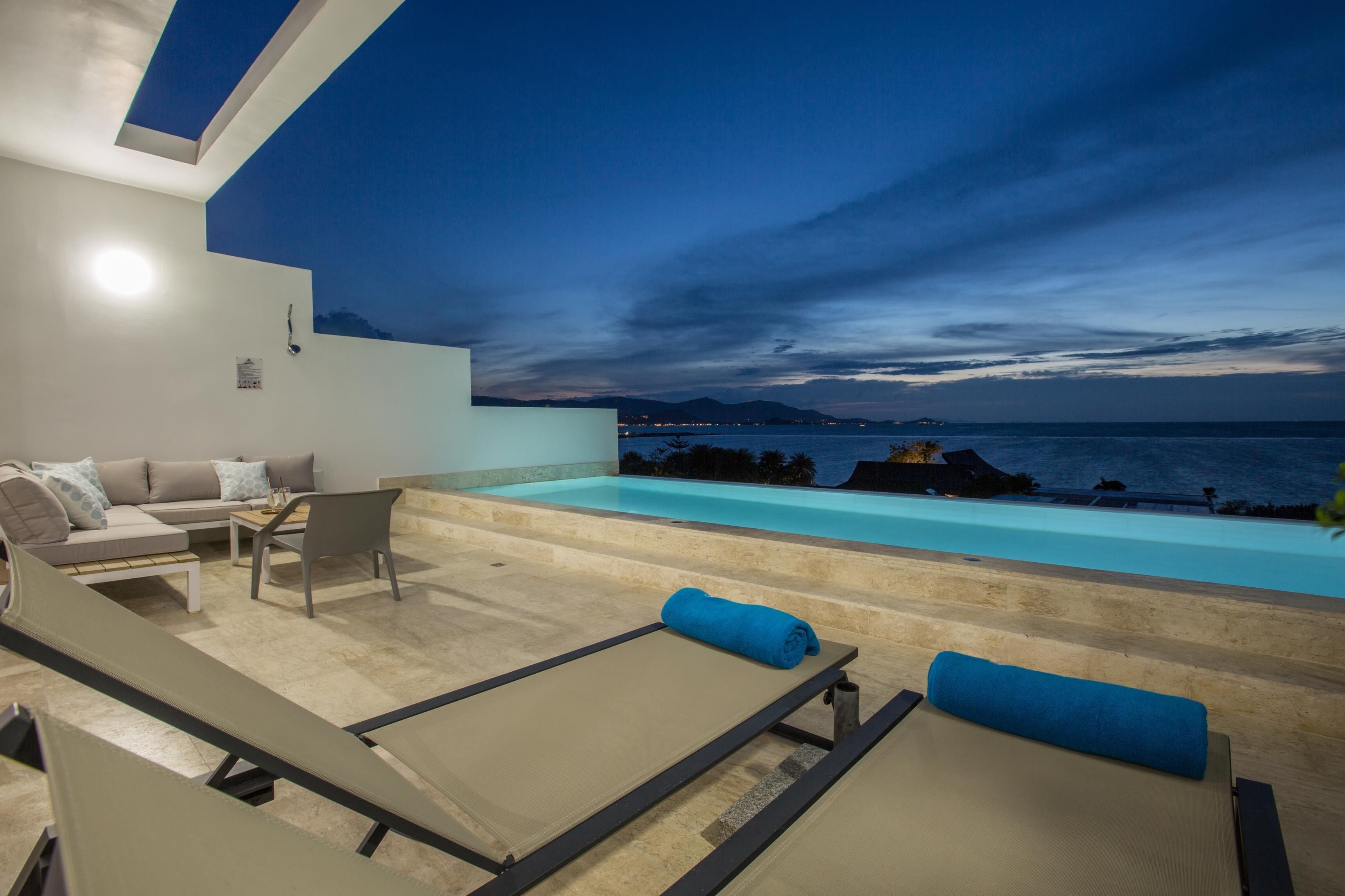 A modern outdoor lounge area with a pool, seating, and a view of the sea at dusk.