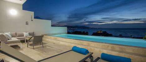 A modern outdoor lounge area with a pool, seating, and a view of the sea at dusk.