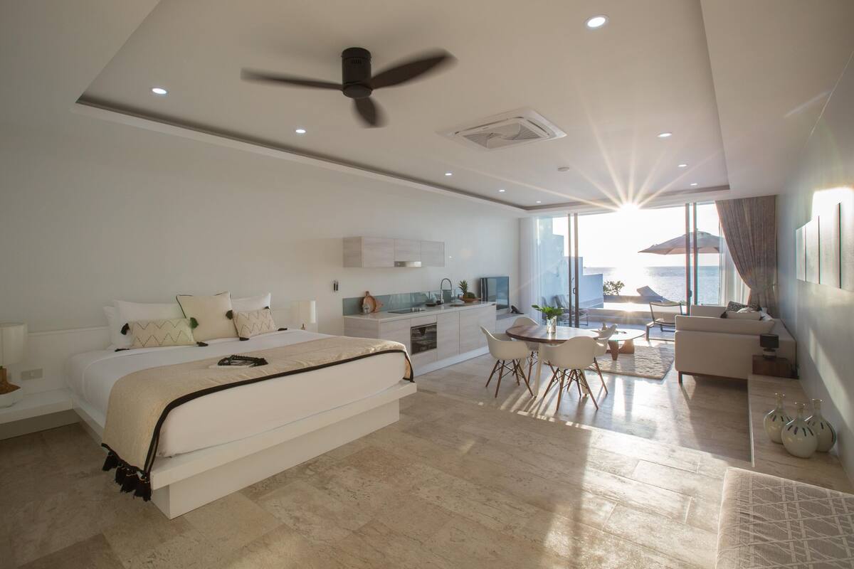 A modern hotel room with a large bed, a dining area, and a view of the sea.