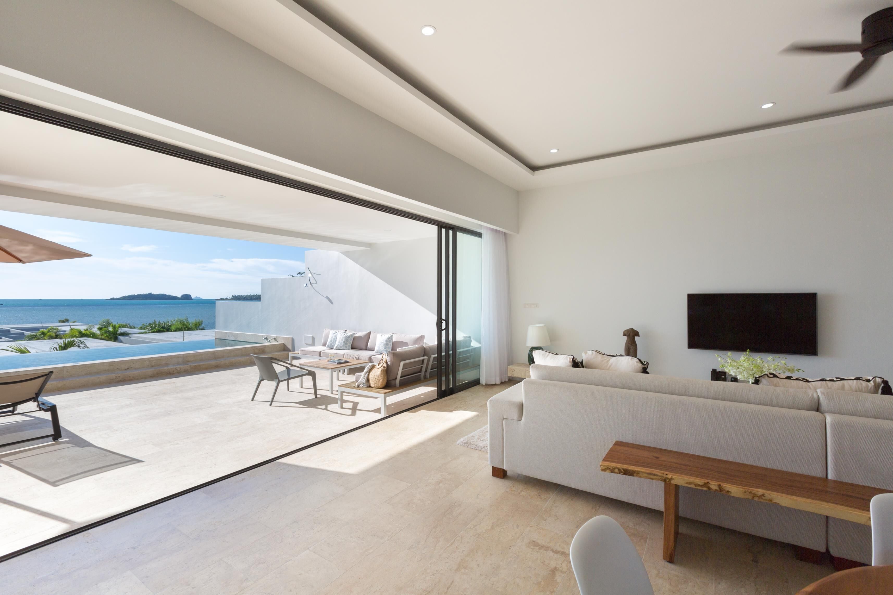 A modern living room with a large sofa, a wooden coffee table, and a TV mounted on the wall. There is a glass door leading to a balcony with a view of the ocean.