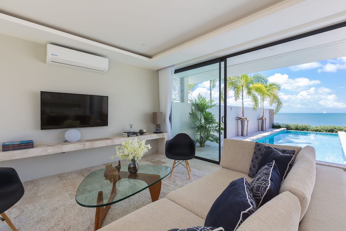 A modern living room with a glass table, a sofa with patterned cushions, a flat-screen TV, and a view of a pool and ocean.