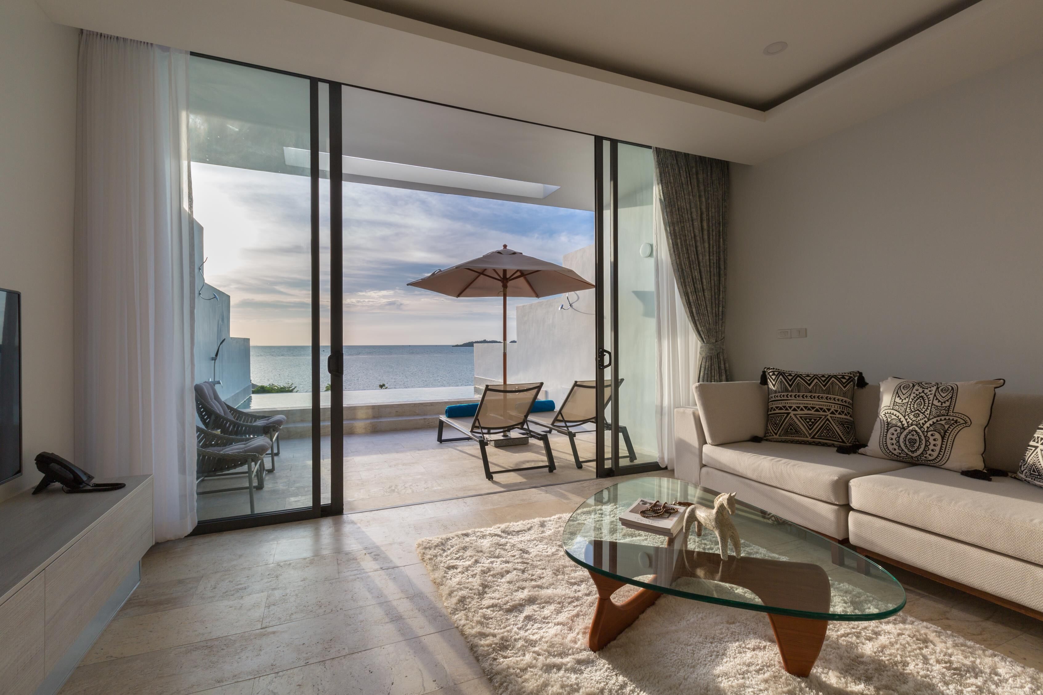 A modern living room with a glass coffee table, a sofa, and a view of the sea through large sliding glass doors.