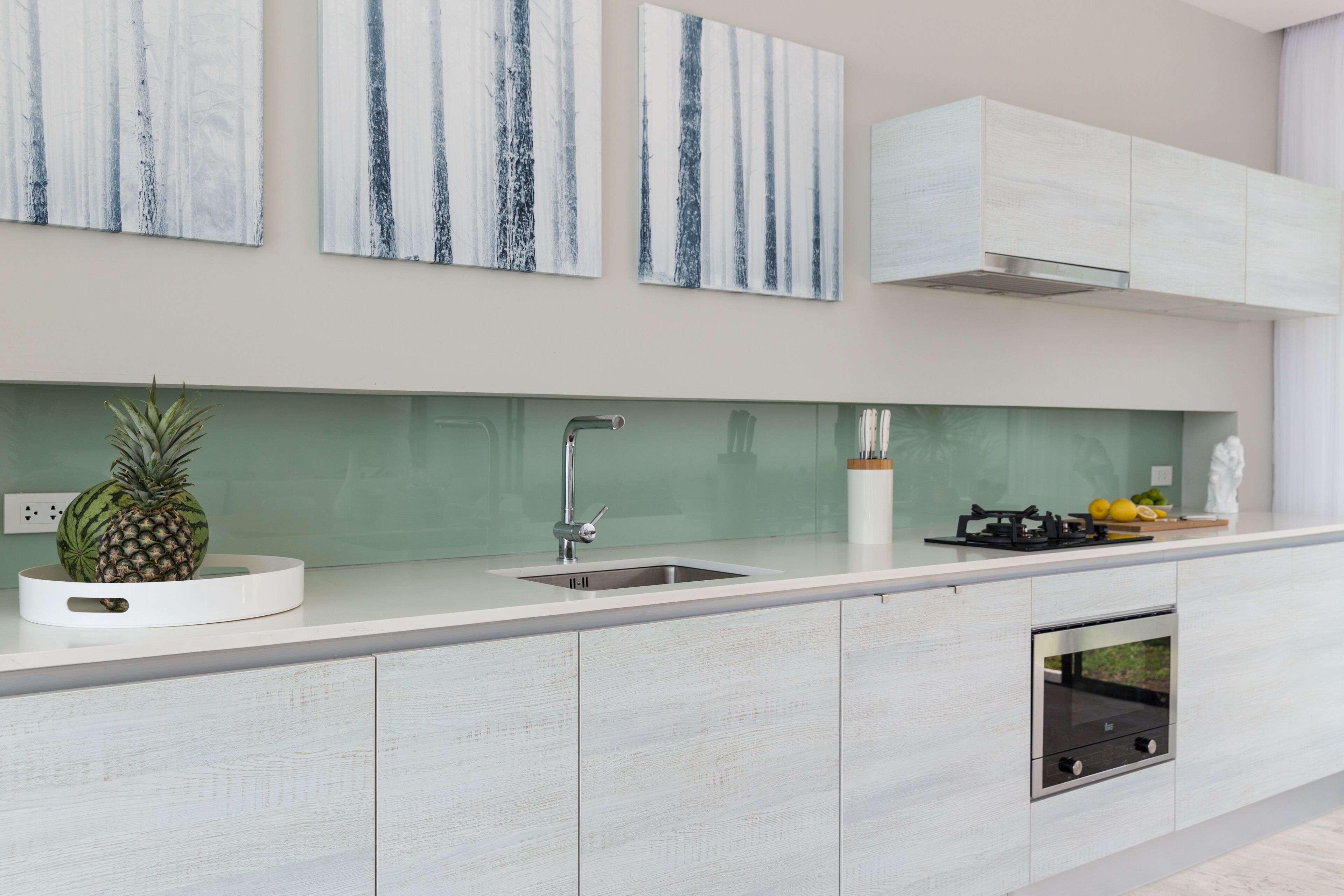 A modern kitchen with a white countertop, stainless steel sink, and built-in oven. There are three abstract paintings on the wall above the counter.