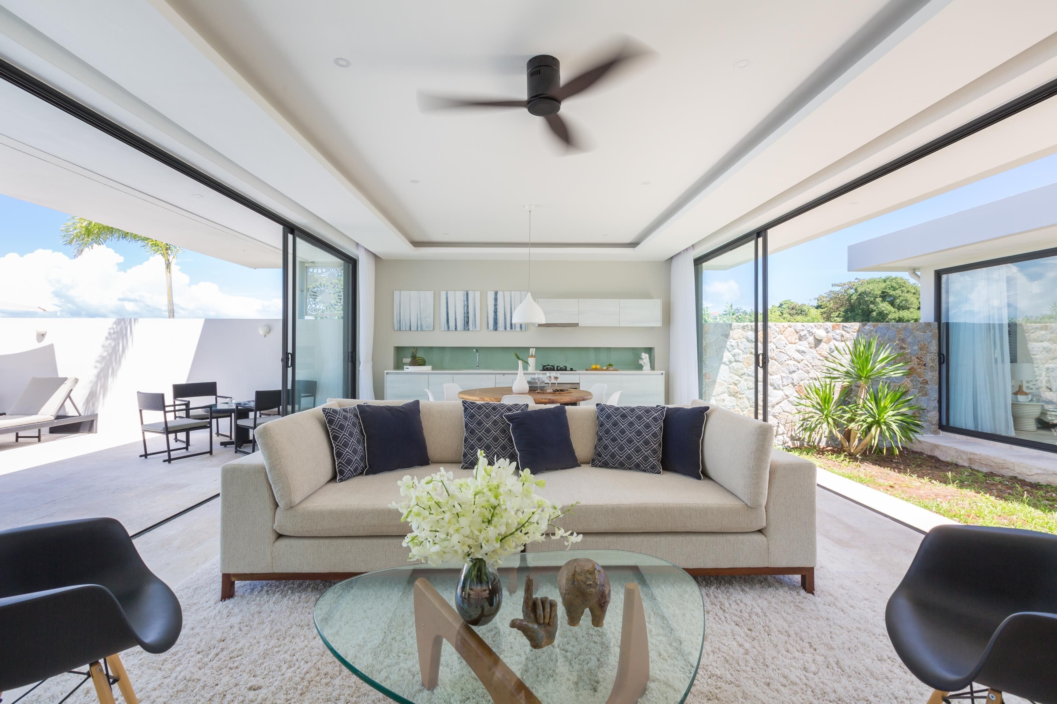 Un salon moderne avec une table basse en verre, un canapé beige agrémenté de coussins à motifs et un ventilateur de plafond.
