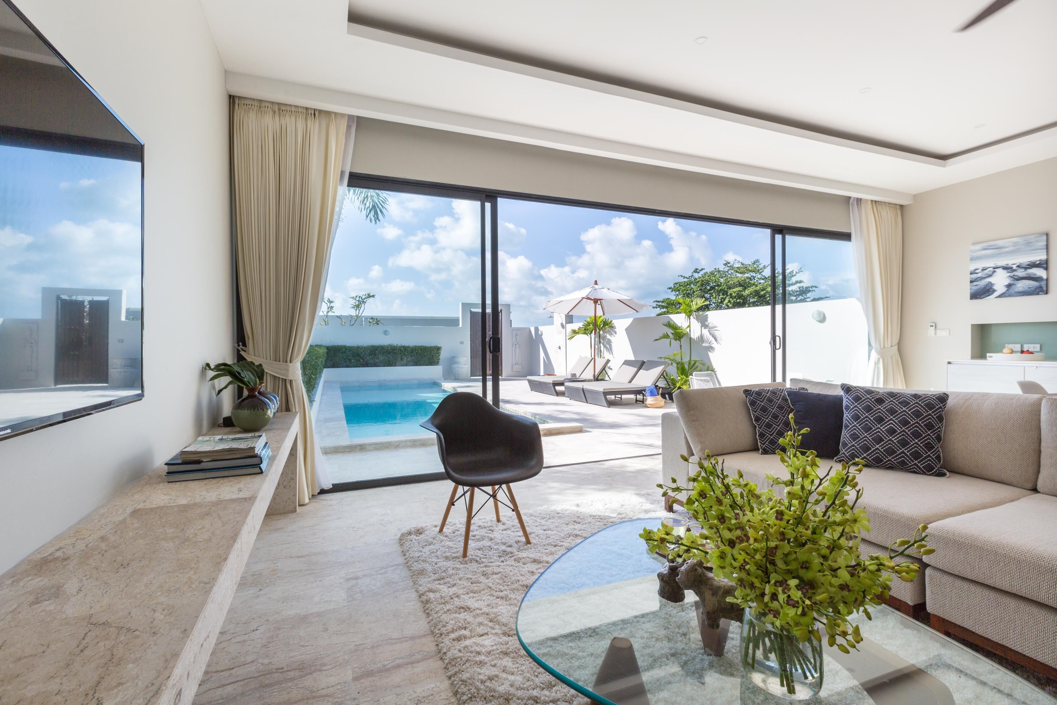 A modern living room with a glass coffee table, a sofa with patterned cushions, a black chair, and a view of a swimming pool and outdoor seating area.