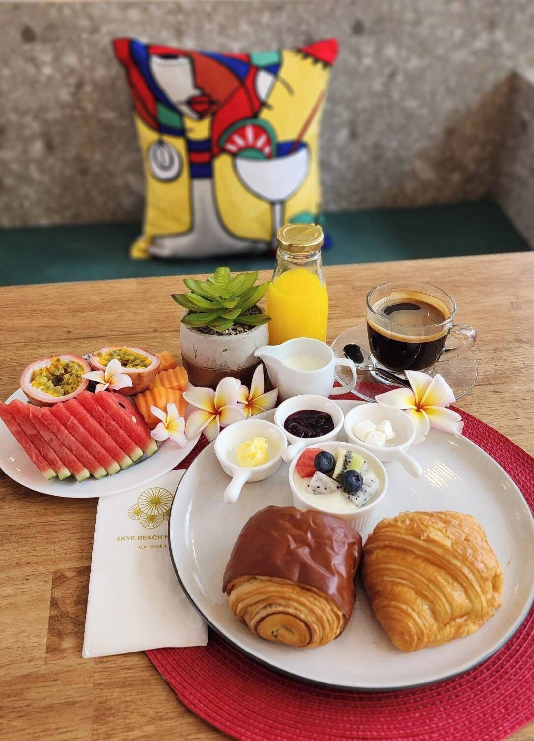 A breakfast setting with a plate of croissants, watermelon slices, and a variety of fruits and condiments.