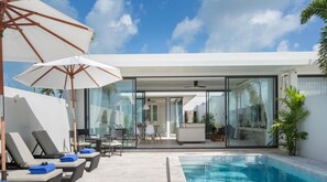 Un área de piscina exterior moderna con sillones de descanso y sombrillas, un espacio habitable con paredes de vidrio y un cielo azul despejado.