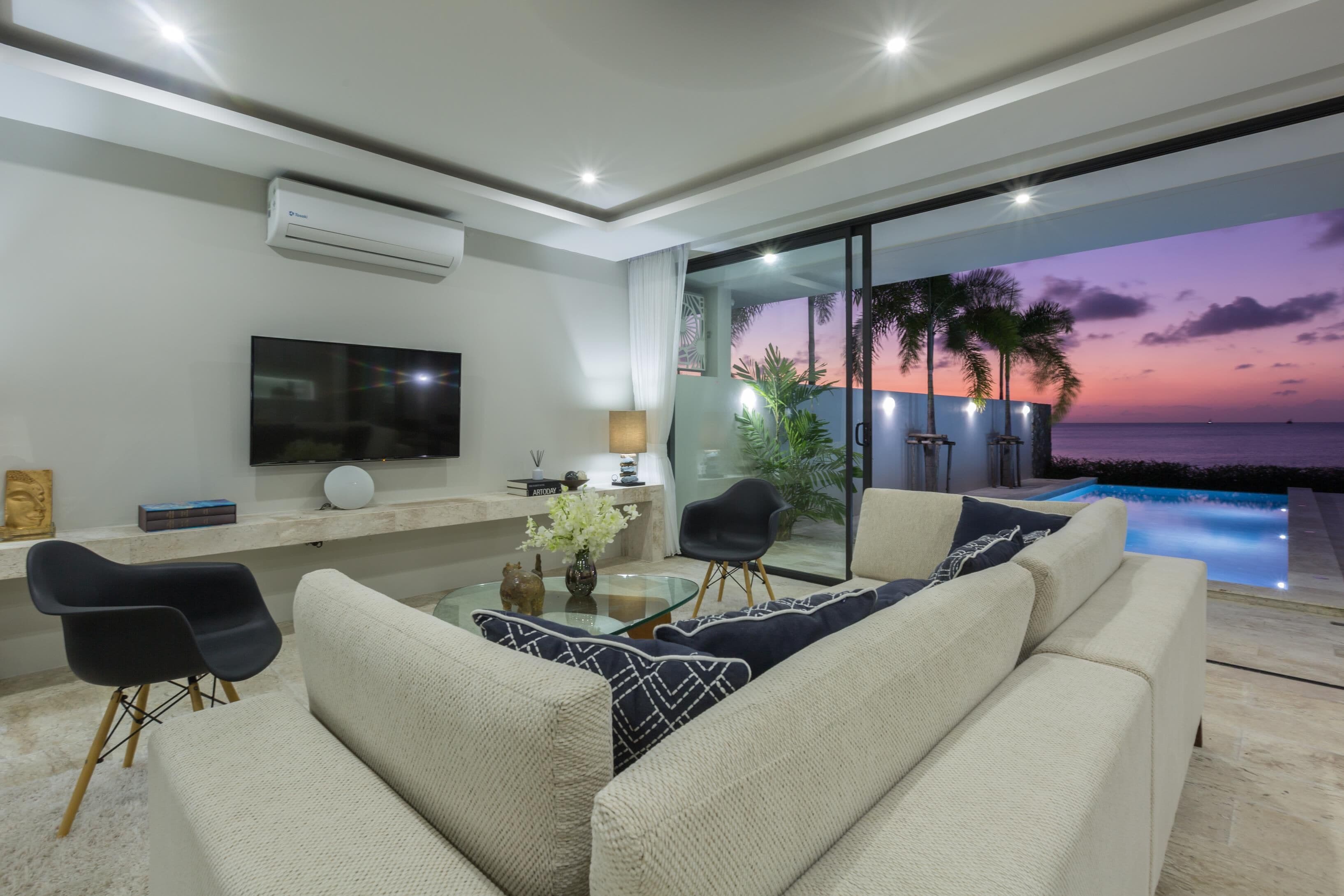 Una sala de estar moderna con un sofá, una mesa de centro y un televisor. Hay un ventanal de vidrio que da a un balcón con piscina y vistas al atardecer.