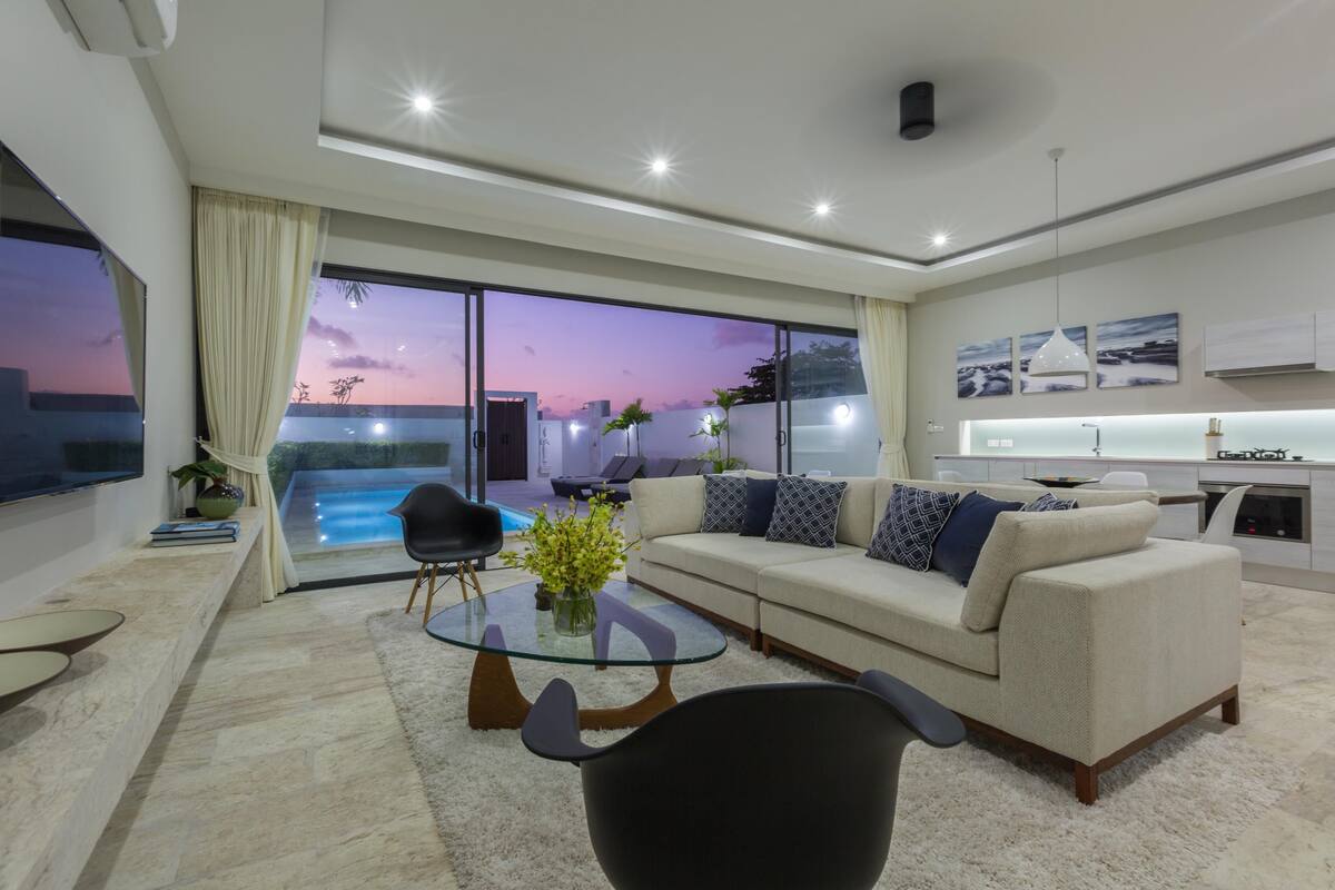 A modern living room with a glass coffee table, a sofa, and a view of an outdoor swimming pool at dusk.