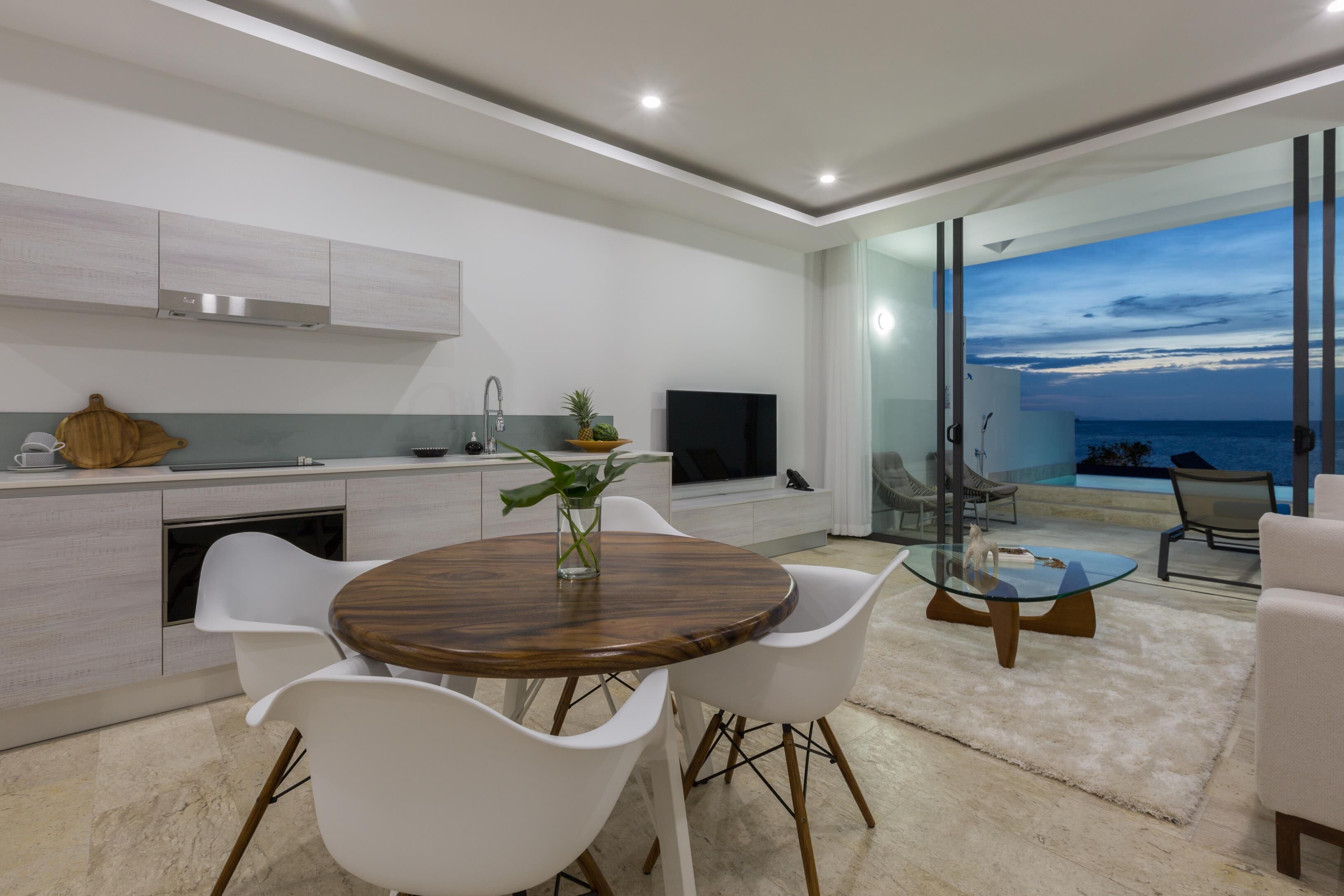 A modern kitchen with white cabinets, a wooden dining table, and white chairs. There is a TV and a plant on the counter.
