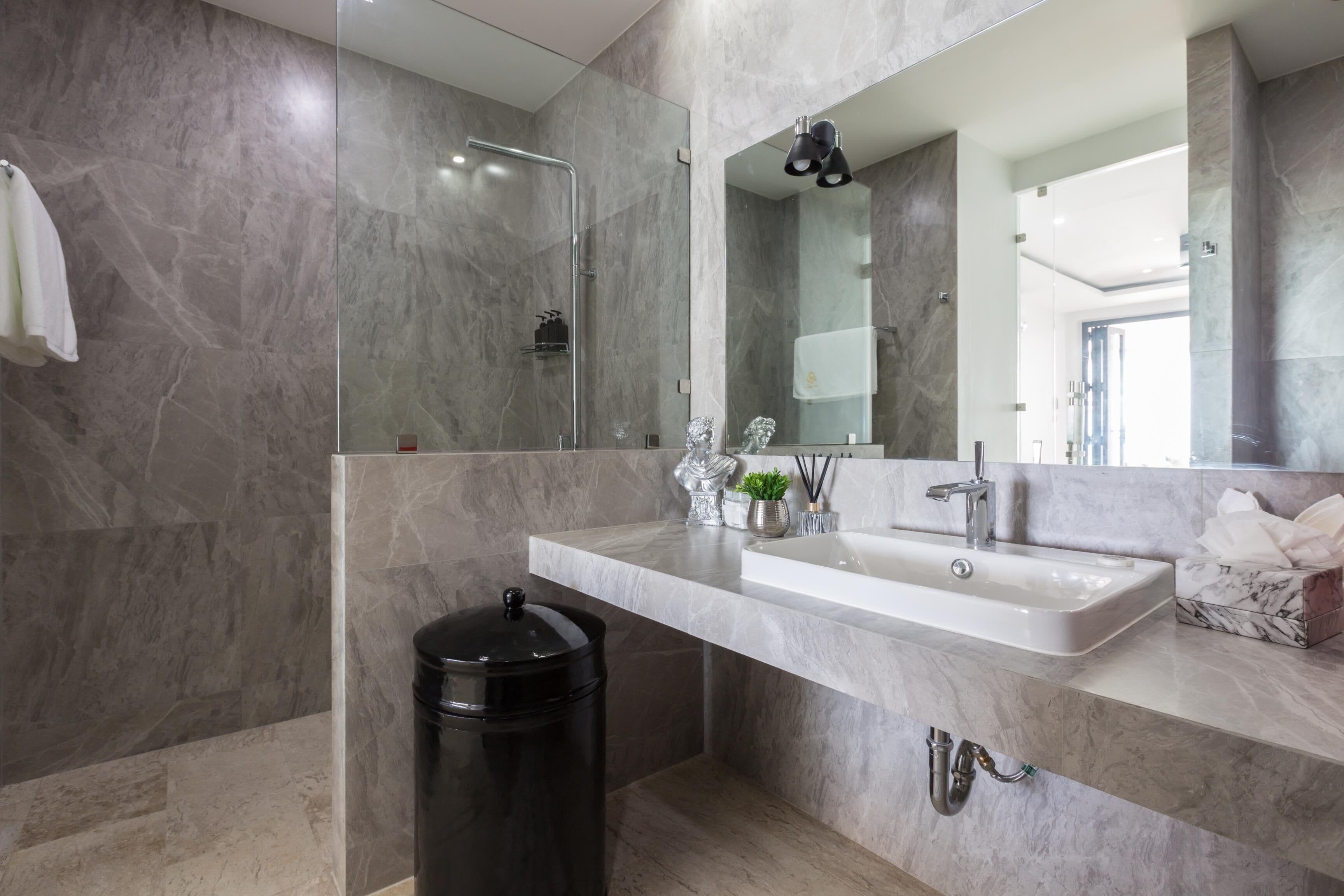A modern bathroom with a marble sink, a glass shower enclosure, and a black trash bin.
