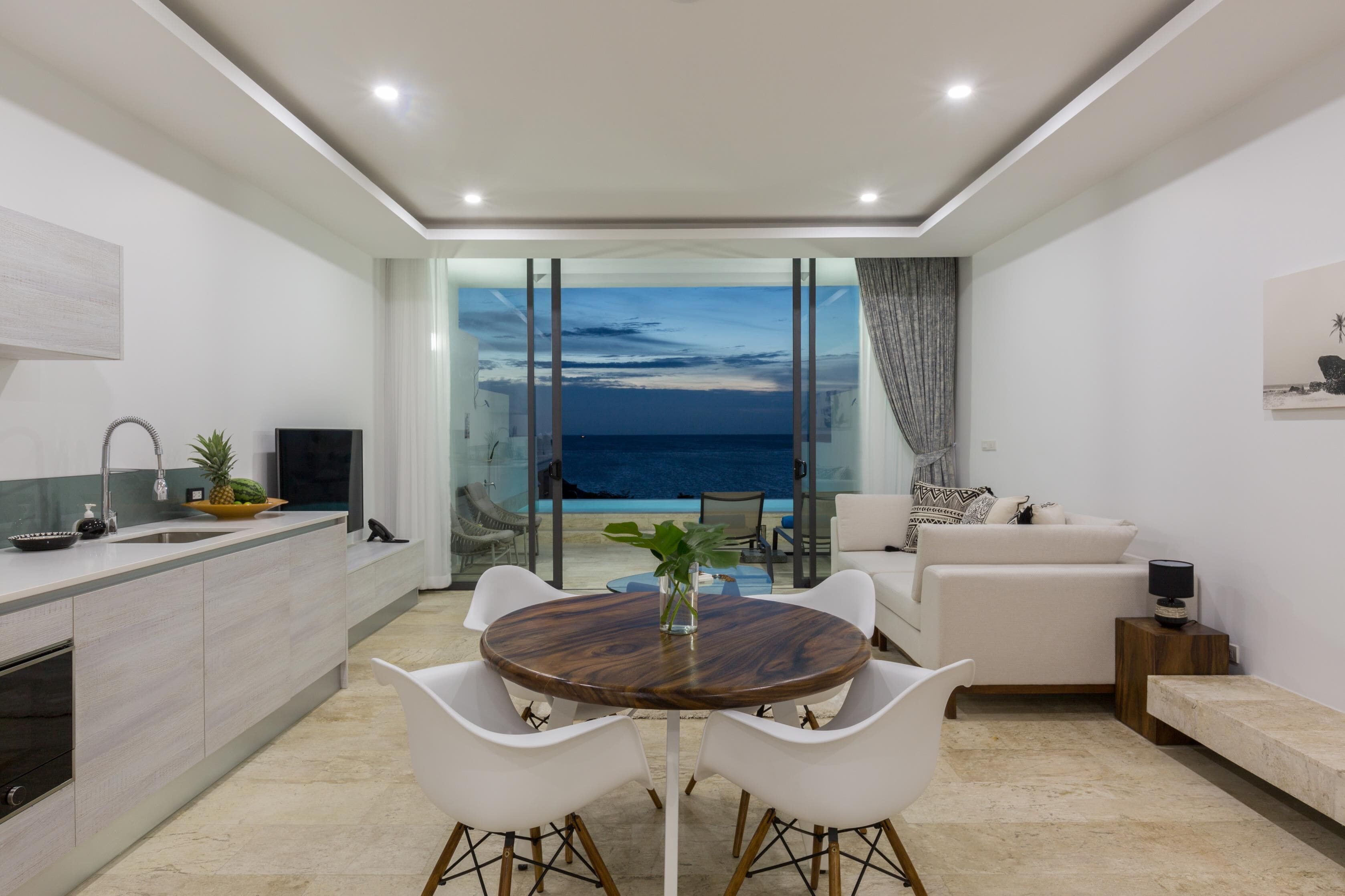 A modern kitchen with a large island, white chairs, and a dining table. There is a view of the ocean through large windows.