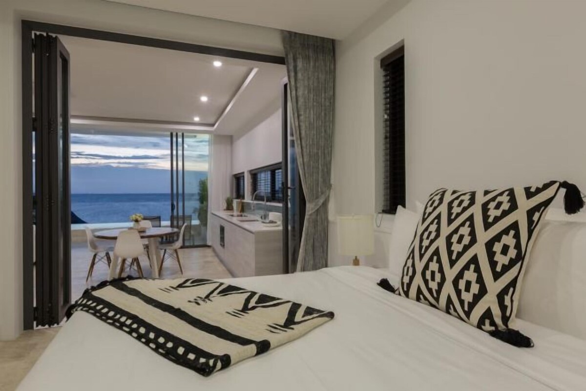 A bedroom with a bed, a bedside table, a window with curtains, and a view of the sea.