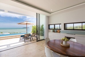 A modern kitchen with a view of the sea, a dining area with a wooden table, and a lounge area with a glass door overlooking the ocean. - Skye Beach Hotel (Koh Samui)