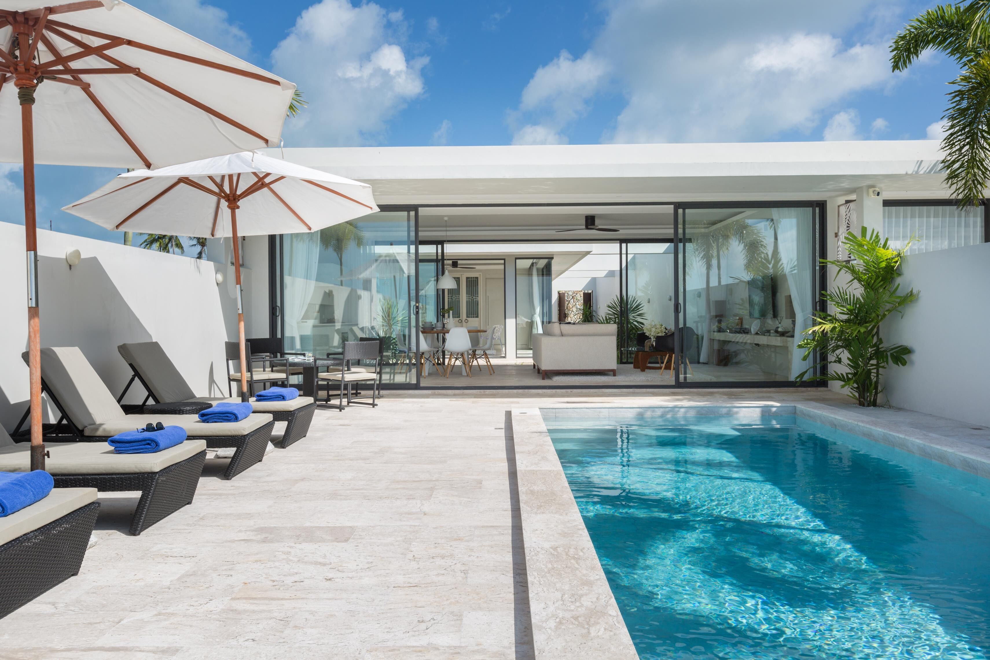 A modern pool area with lounge chairs and umbrellas, a glass-enclosed patio, and a clear blue sky.