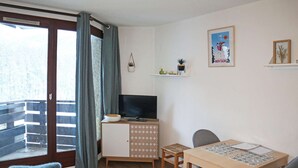 Une petite pièce bien éclairée, avec un plancher en bois, un téléviseur posé sur un meuble, une table à manger entourée de chaises et un balcon d’où l’on peut admirer les arbres.