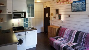 Une cuisine compacte équipée d’un four à micro-ondes, d’un four et d’un lave-linge. On y trouve un meuble en bois et un canapé à rayures. Une pancarte indique : « Amour Flowers ».