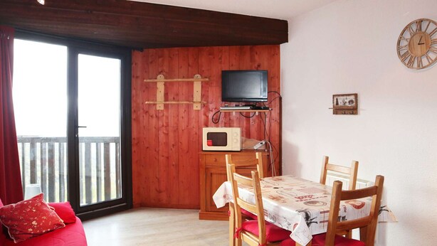 A cozy room with a wooden wall, a television mounted on a shelf, a microwave, a dining table with chairs, and a red sofa.