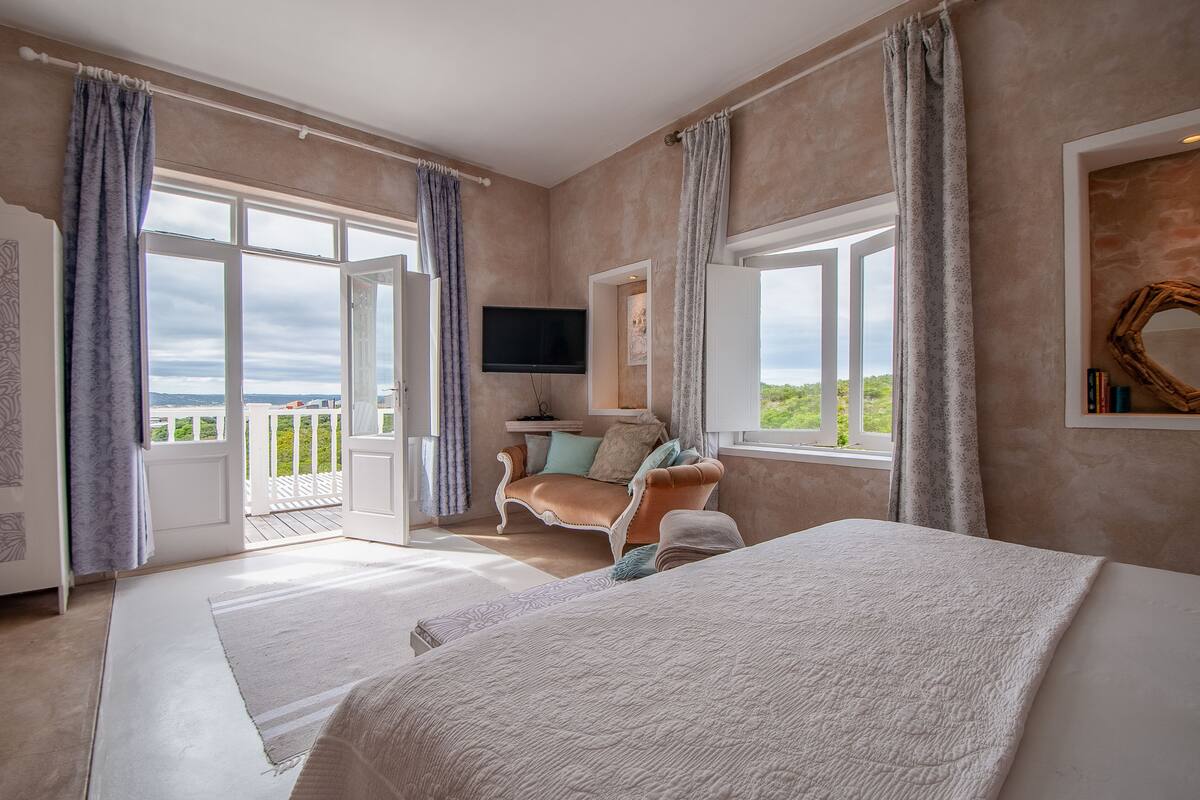 A bedroom with a large bed, a sofa, a TV, and a balcony with curtains.