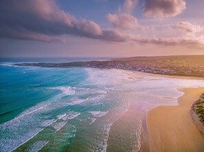 Une vue aérienne d’une zone côtière avec une plage, l’océan et, au loin, une étendue terrestre parsemée de bâtiments.