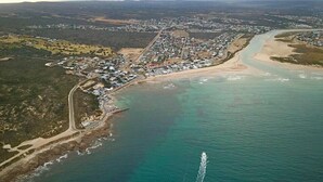 An aerial view of a coastal town with a prominent road leading to the beach, a clear blue sea, and a visible boat wake. - Charlotte House (Still Bay)