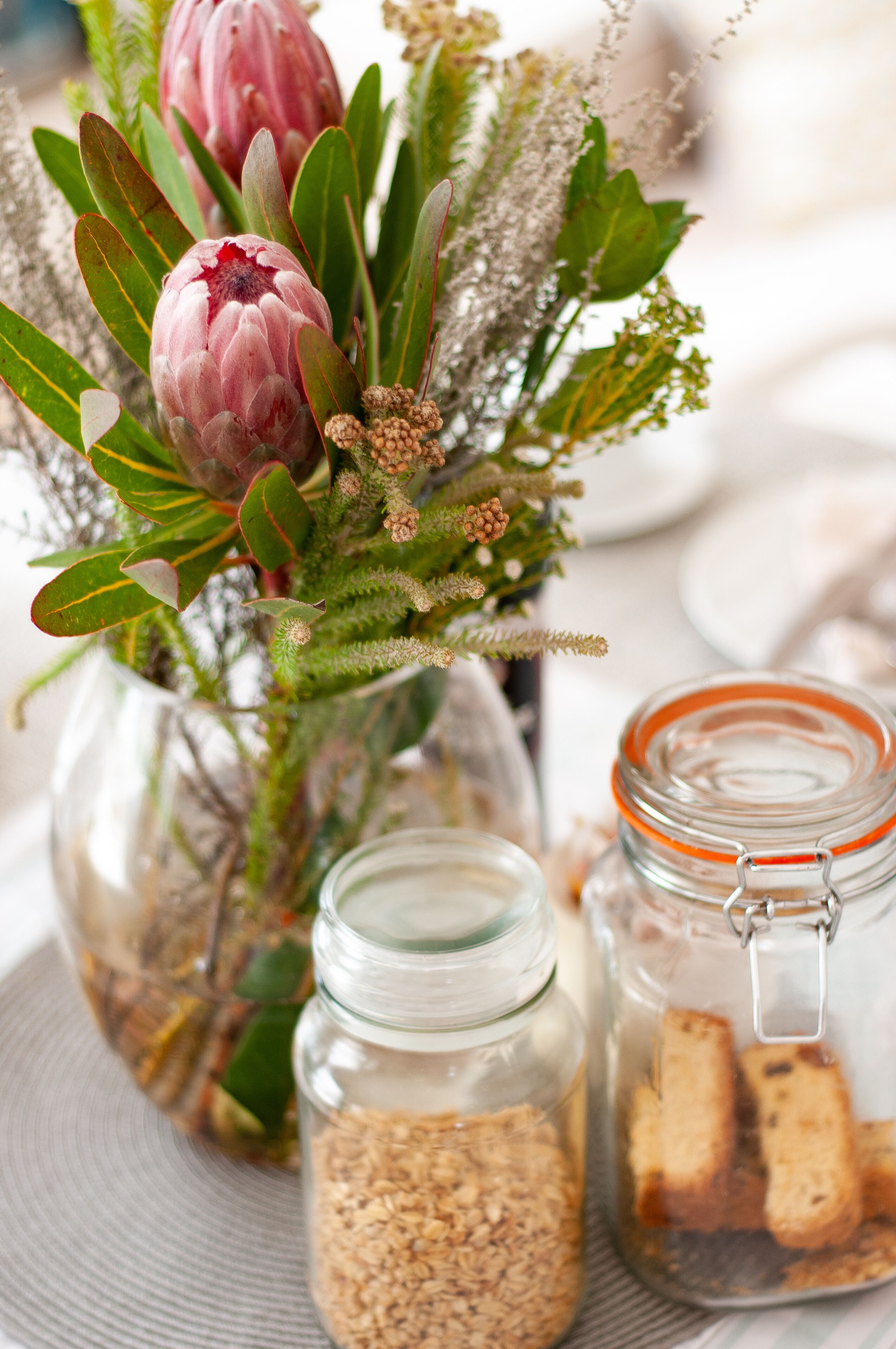 A close-up of a glass jar filled with oats, a glass jar with a metal clasp containing bread, and a glass vase with flowers and greenery.