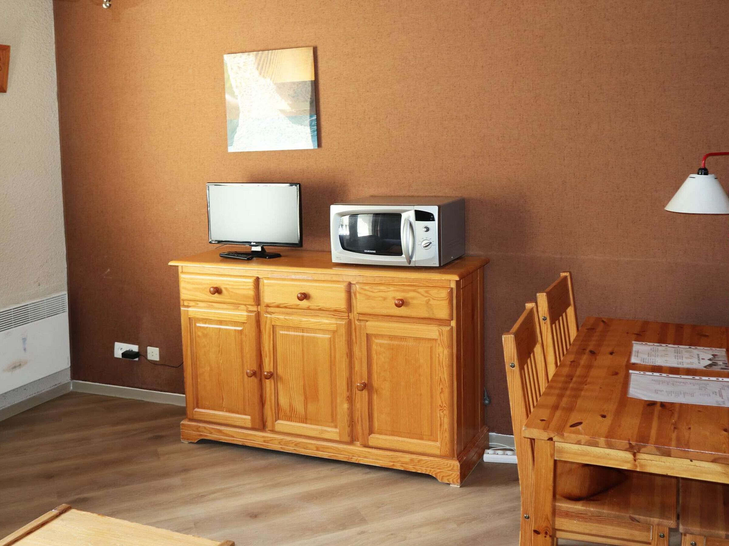 Un meuble en bois sur lequel sont posés un téléviseur et un four à micro-ondes, une table à manger avec des chaises, et un luminaire fixé au mur.