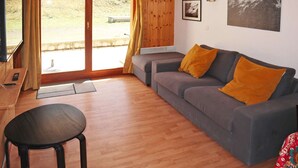 Sala de estar com sofá cinza, uma cadeira preta e uma mesa de madeira com um controle remoto. Há duas janelas com cortinas, um aquecedor fixado na parede e duas fotos emolduradas.