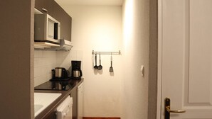 Cozinha pequena com micro-ondas, cafeteira e máquina de lavar louça. Possui um balcão compacto e uma porta com puxador dourado.