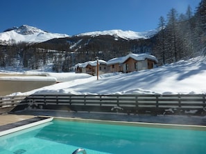A pool area with a view of snow-covered mountains and wooden chalets. - Charming Chalet for 6 in Les Orres 1800 - Pool, Sauna, Hammam, Balcony, Linens Included (Les Orres)