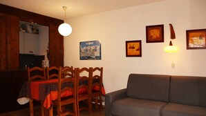 Ett vardagsrum med en grå soffa, ett matbord i trä med stolar och ett soffbord i trä. Det finns vägglampor och inramade tavlor på väggen.