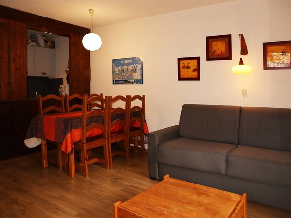 A living room with a grey sofa, wooden dining table with chairs, and a wooden coffee table. There are wall-mounted lights and framed pictures on the wall.