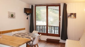 Una habitación pequeña y acogedora con una mesa de madera y sillas, un televisor en la pared y un balcón con vistas a un paisaje montañoso.