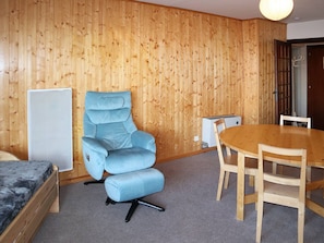 A room with a blue reclining chair, a wooden dining table with four chairs, a white cabinet, and a bed with a dark headboard. - 2-Room Apartment for 6 - Centrally Located, Pets Allowed, Parking (Les Orres)