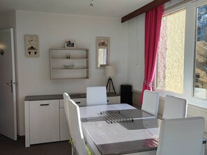 Een compacte hotelkamer met een kitchenette, een eettafel met stoelen, een klein plankje met decoratieve spullen, een raam met gordijnen en een deur.