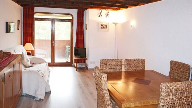A cozy room with a wooden dining table, wicker chairs, a television, and a bed with a red curtain. - Central Apartment, Ski-in/Ski-out, Sleeps 6, Balcony, South-Facing (Les Orres)