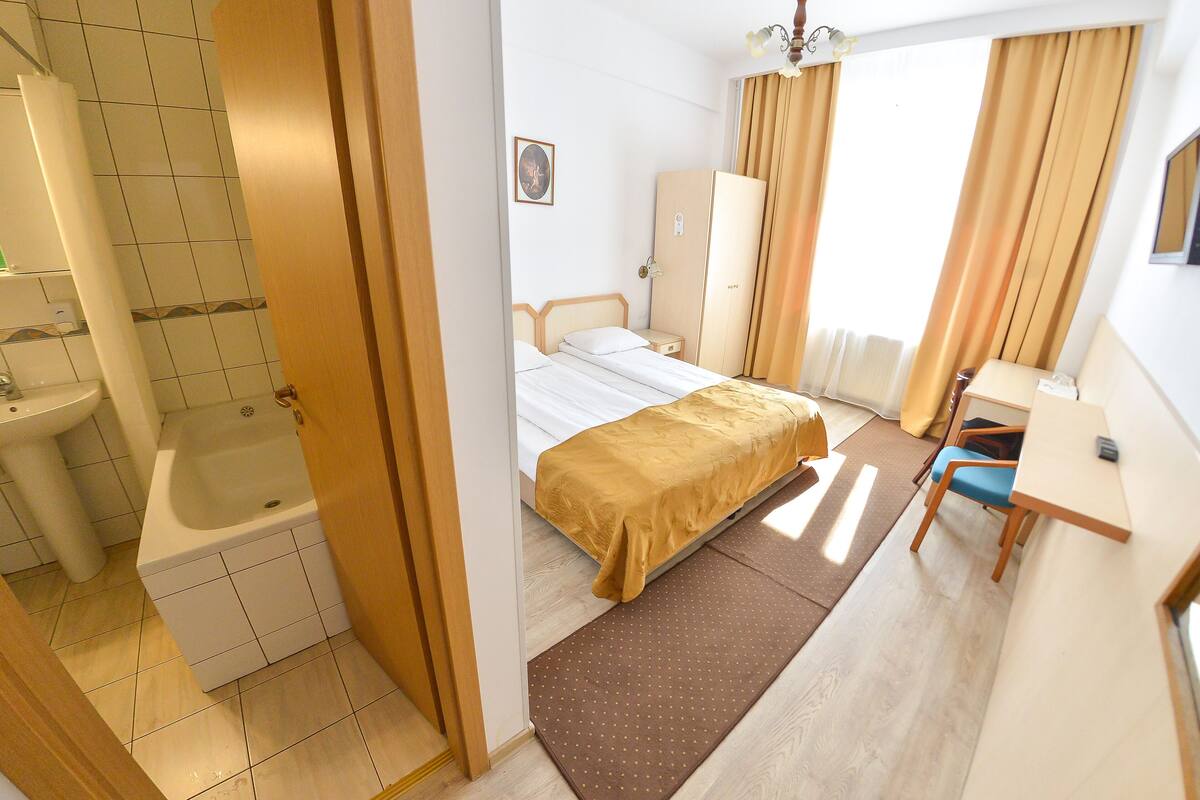 Ein Hotelzimmer mit Bett, Schreibtisch, Stuhl, Kleiderschrank, Bad mit Waschbecken und Badewanne sowie einem Fenster mit Vorhängen.