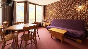 Una habitación con una mesa de comedor de madera, sillas, un sofá púrpura, un banco de madera, un televisor fijado en una pared de madera y un papel tapiz estampado.