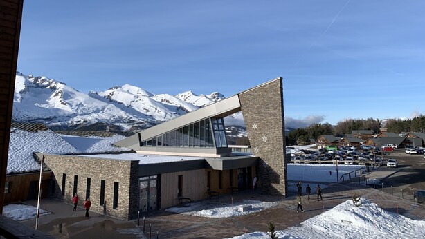 Un edificio moderno con tejado inclinado y ventanas grandes, con montañas nevadas al fondo.