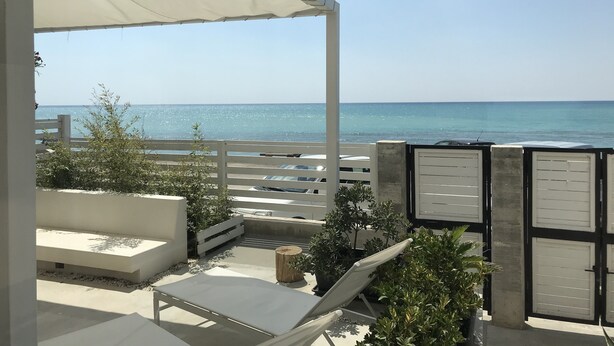 Un'area relax all'aperto coperta con vista sul mare, arredi bianchi e una recinzione in legno.