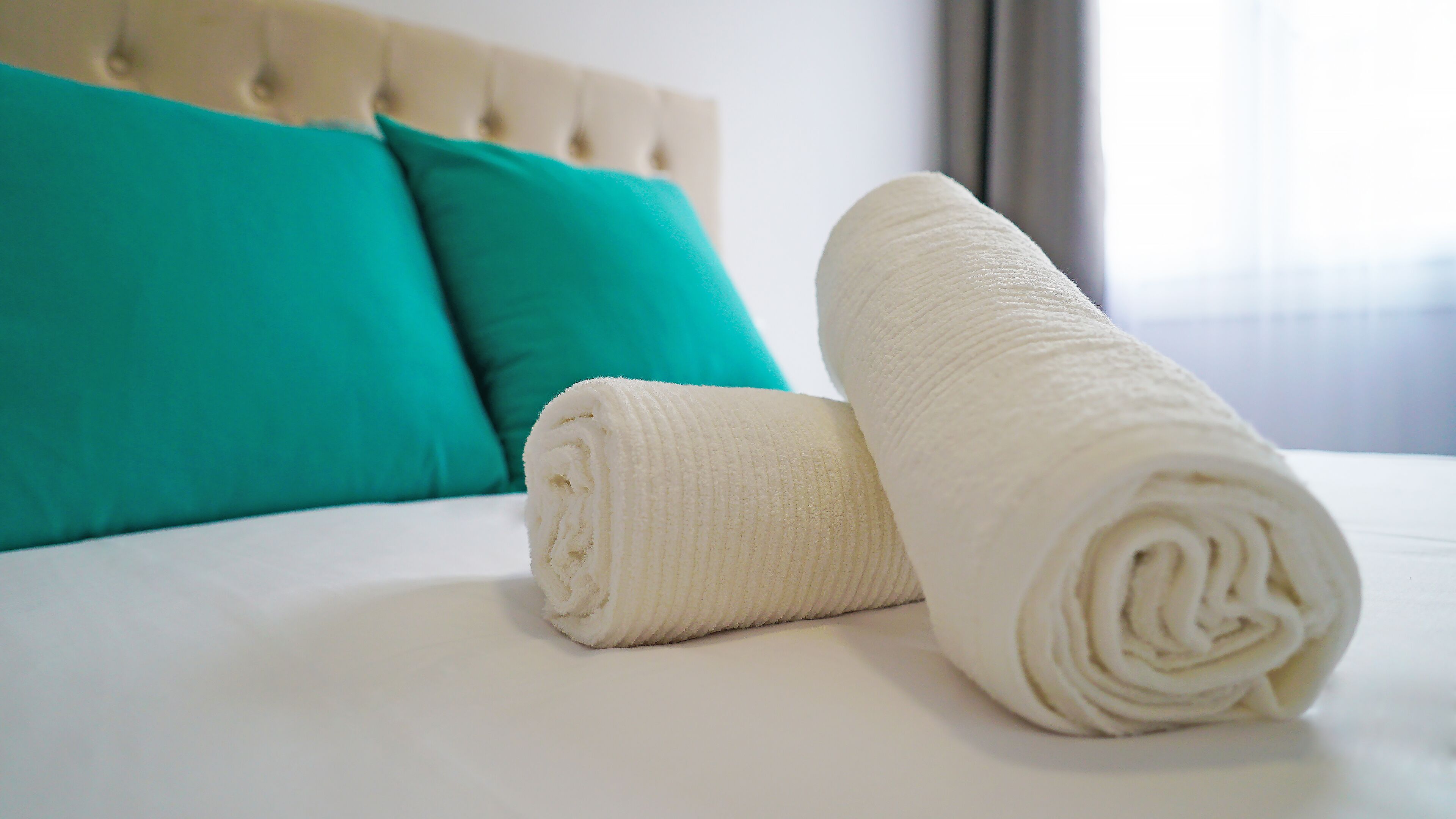Una cama bien hecha con sábanas blancas, dos toallas enrolladas, un cojín color turquesa y un cabecero capitonado.
