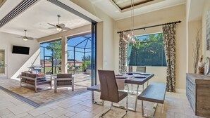 A spacious living area with a dining table, chairs, and a sofa. There is a large window with curtains, a ceiling fan, and a TV mounted on the wall.