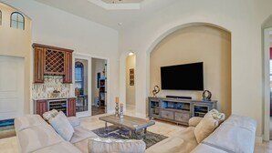A spacious living room with a large sofa set, a flat-screen TV, and a kitchen area visible through an archway.