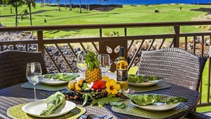 Une table dressée avec des assiettes, des verres, une bouteille et des fruits tropicaux, donnant sur un terrain de golf et l’océan.