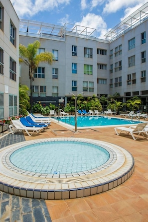 Un área de piscina en la azotea con sillones de descanso y un jacuzzi circular, rodeada por un edificio con múltiples ventanas y balcones.